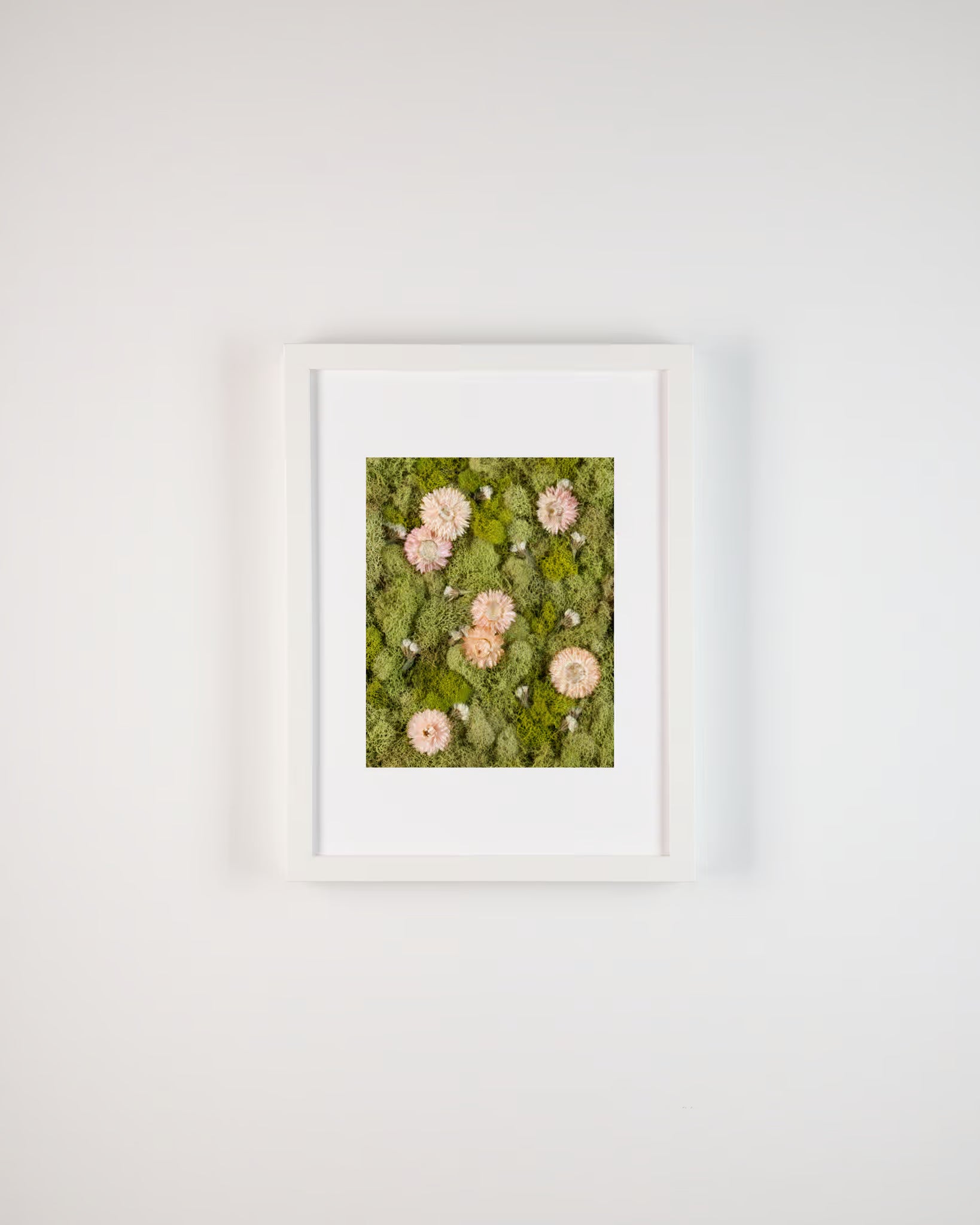 Framed artwork of pink flowers on green moss against a white wall